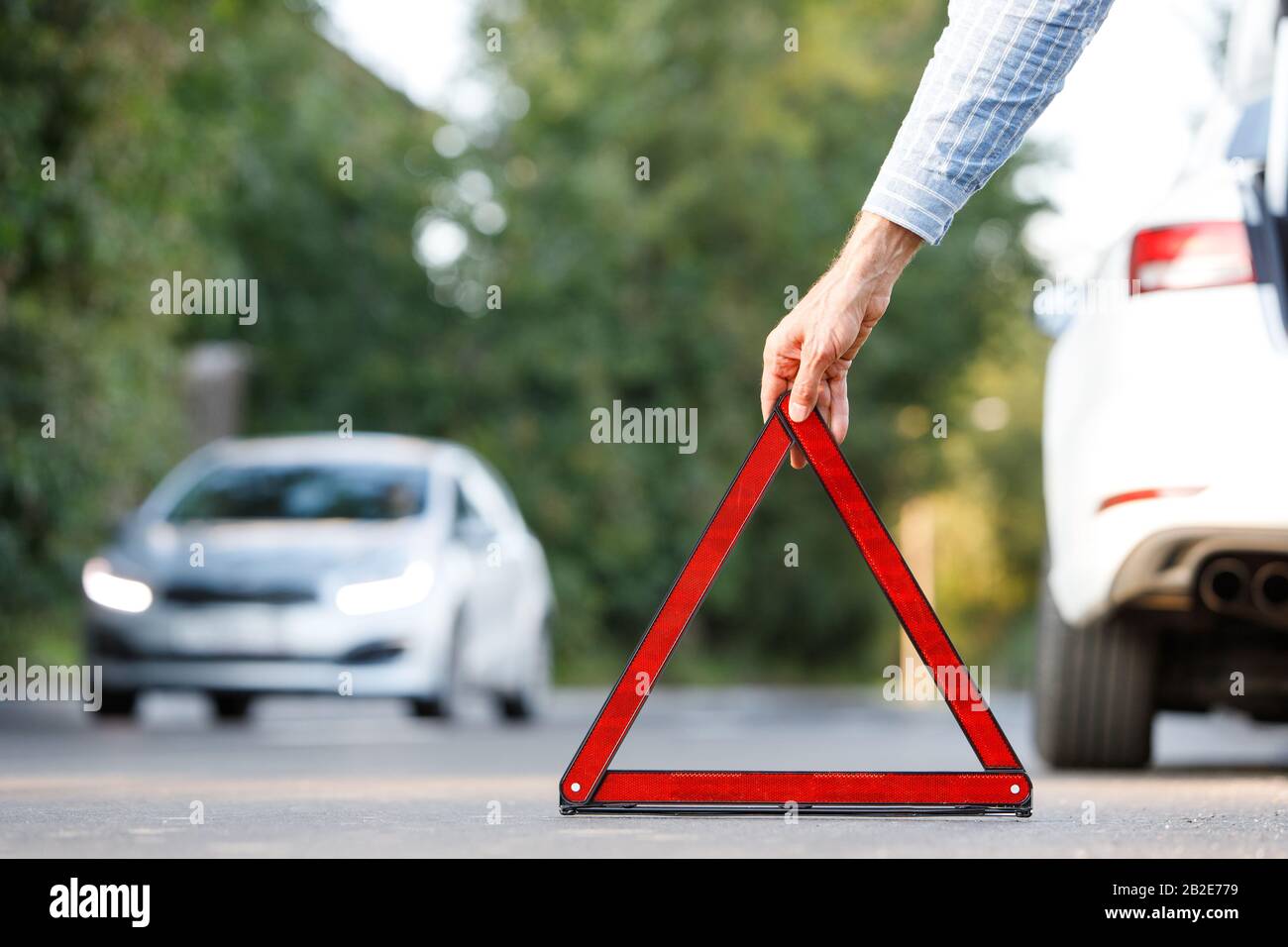 Close up of man driver putting red warning triangle/emergency stop sign behind his broken car on the side of the road, copy space and blurred car on b Stock Photo