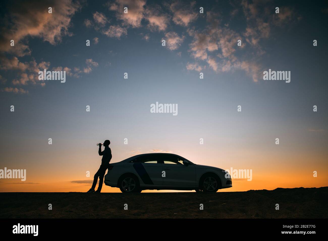 Silhouette of man driver relaxing after a ride, standing next to his car and drinking water from a bottle, side view. Sunset time. Stock Photo