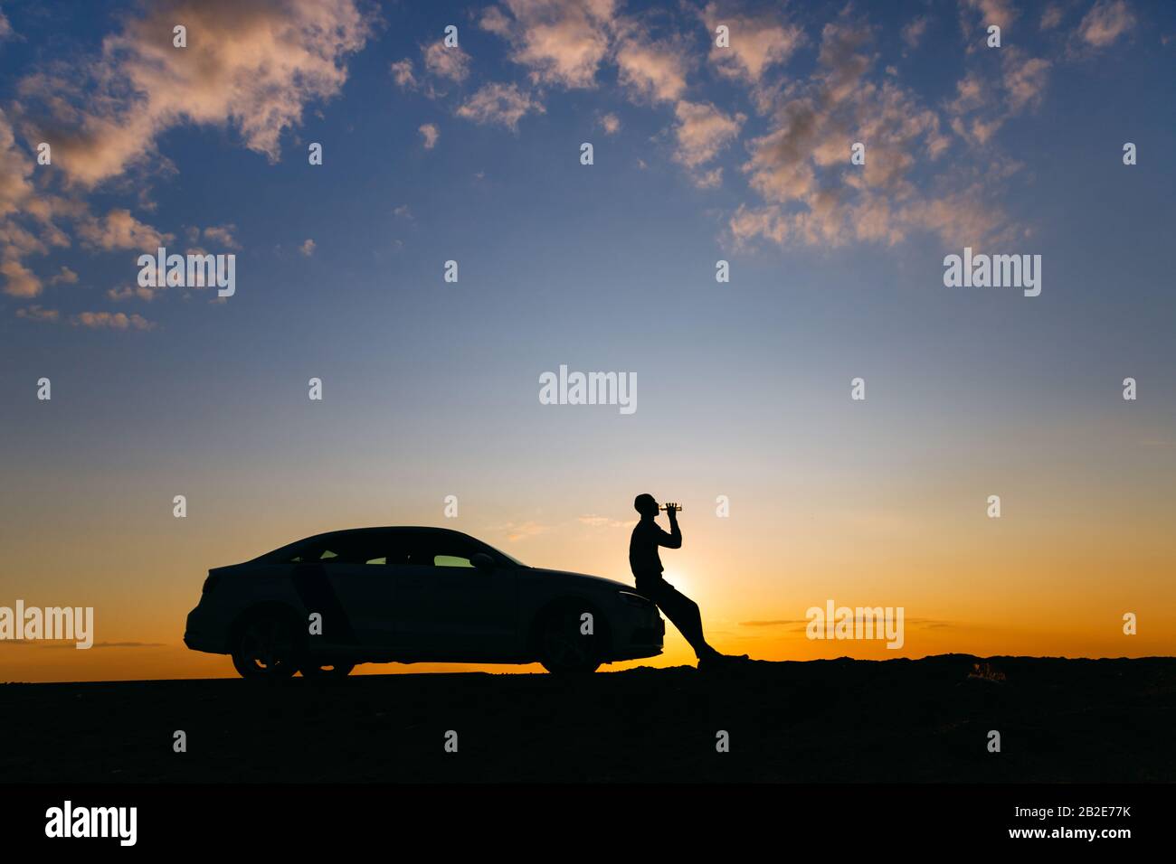 Silhouette of man driver relaxing after a ride, sitting on the hood of his car and drinking water from a bottle, side view. Sunset time. Stock Photo