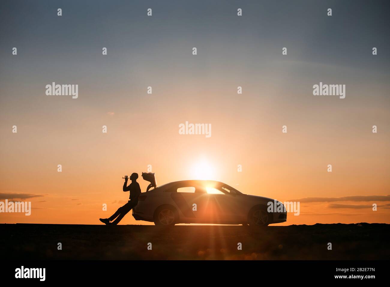 Silhouette of man driver relaxing after a ride, sitting on the trunk of his car and drinking water from a bottle, side view. Sunset time. Stock Photo