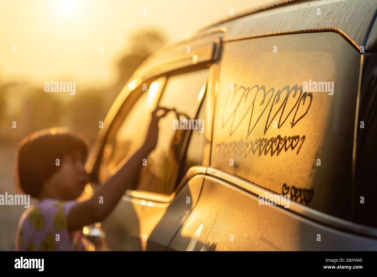 Asian little girl writing or drawing heart symbol on wet mirror of her father SUV car in morning for love concept. Focus on mirror Stock Photo