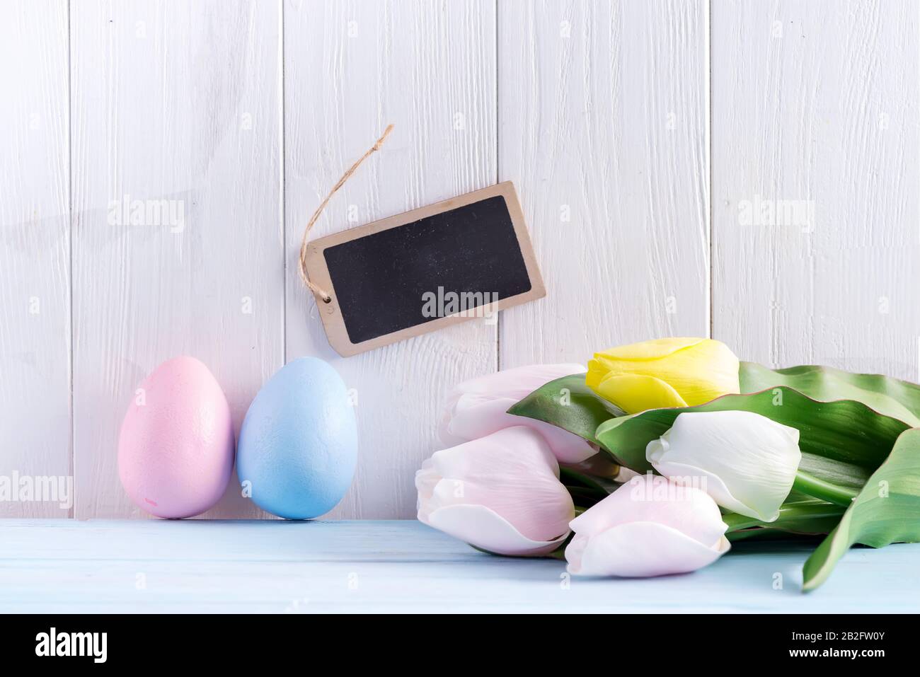 Happy Easter card from craft painted eggs, chalkboard for message and fresh tulips against light grey wooden background. Stock Photo
