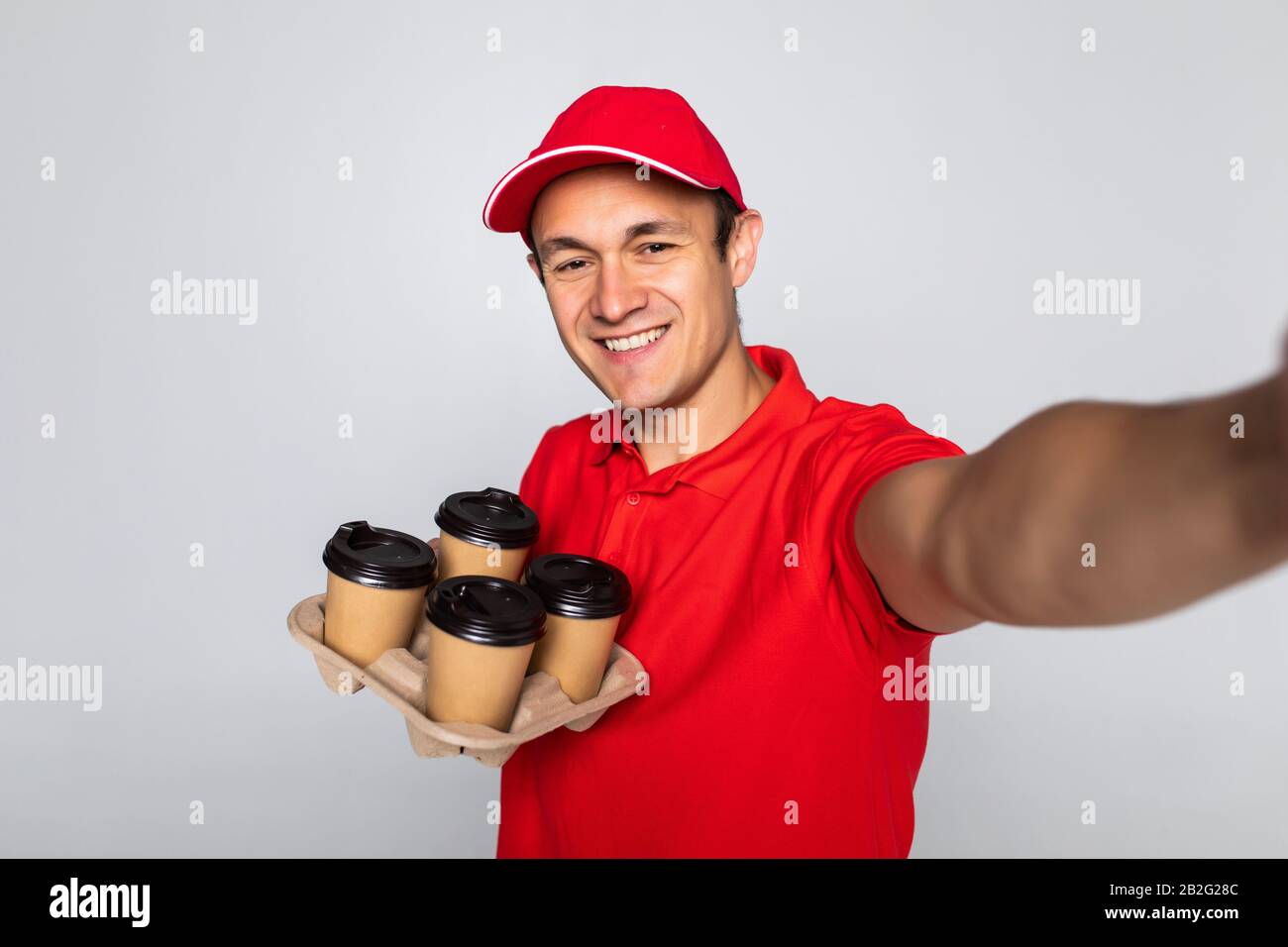Happy man from delivery service in red t-shirt and cap giving food order and holding coffee boxes over white background Stock Photo