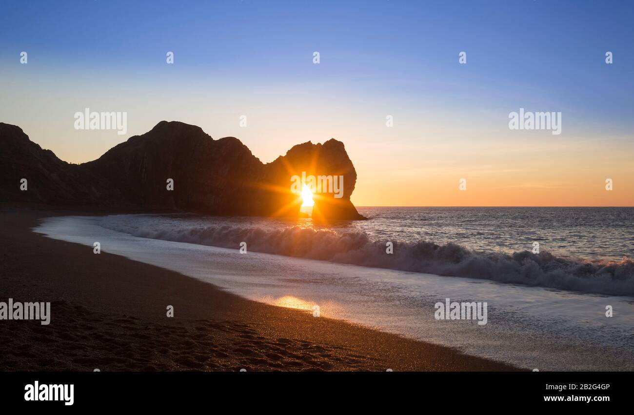 Sunrise through Durdle Door arch and waves breaking on beach, Jurassic Coast, Dorset, England, UK Stock Photo