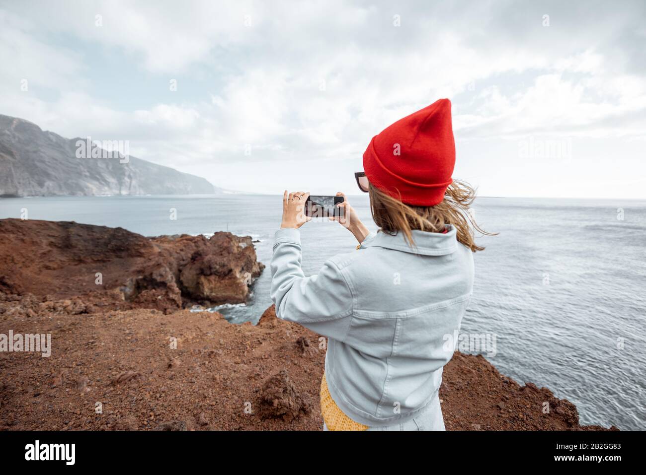 Young woman in red hat enjoying a trip on a rocky ocean coast, photographing on phone breathtaking landscapes. Traveling on Tenerife island, Spain Stock Photo