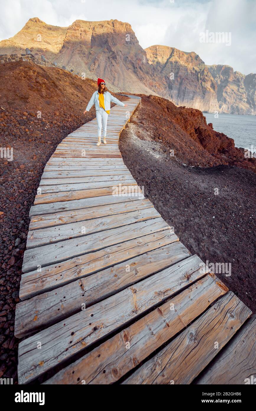 Woman walking on the picturesque wooden pathway through the rocky land with mountains on the background. Traveling on the north-west cape of Tenerife island, Spain Stock Photo