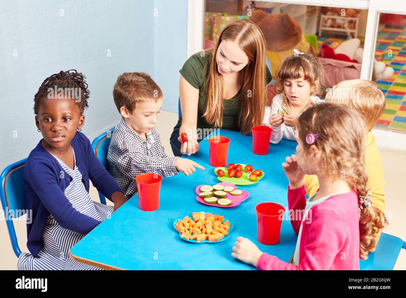 Children in kindergarten eat vegetables as a snack at the table with a ...