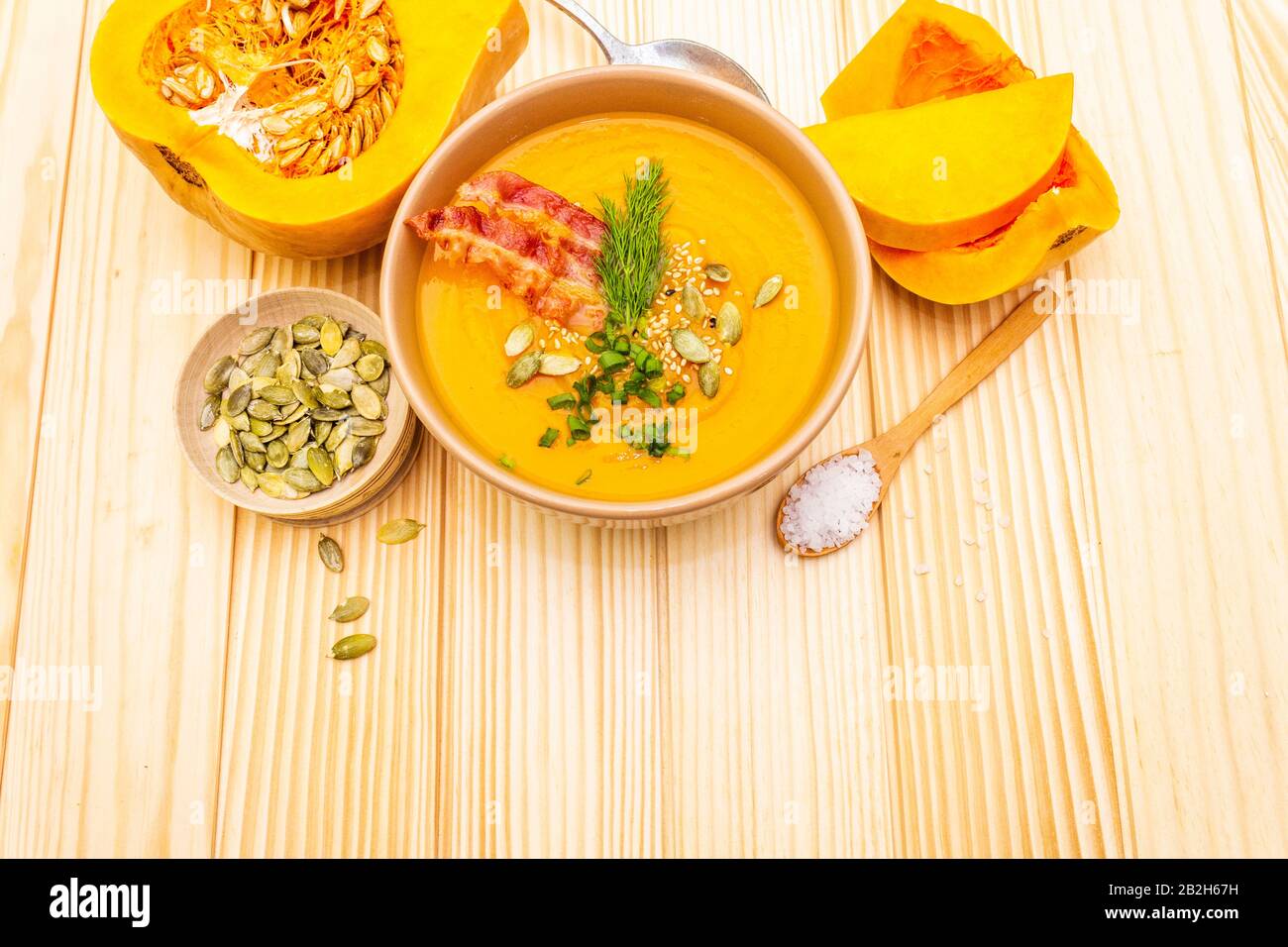 Homemade autumn hot pumpkin cream soup with smocked bacon and seeds. Raw pumpkin, fresh chives, dill, salt in spoon on wooden background Stock Photo