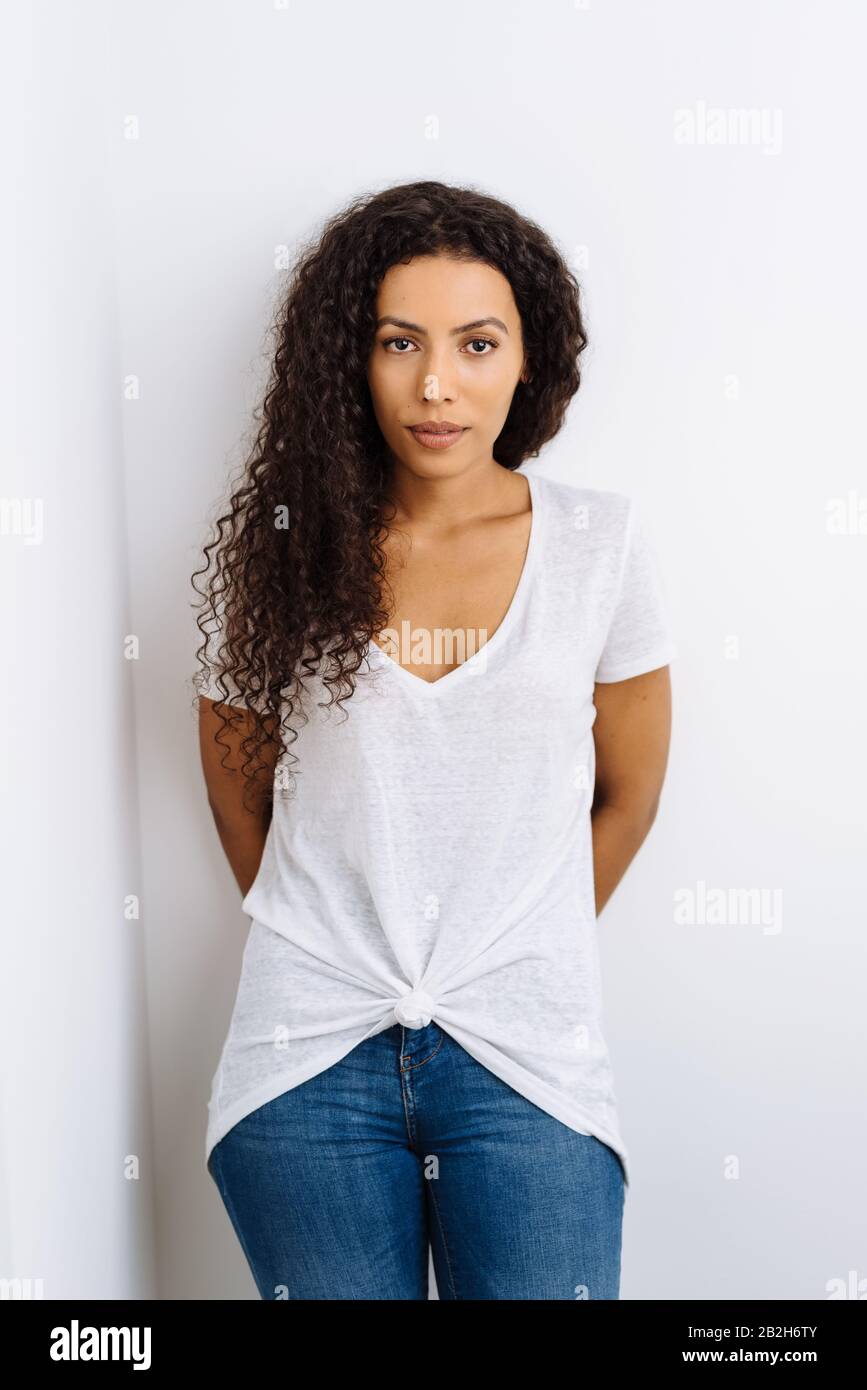 Serious young African woman in jeans standing staring intently at the camera with her hands behind her back against an interior white wall Stock Photo