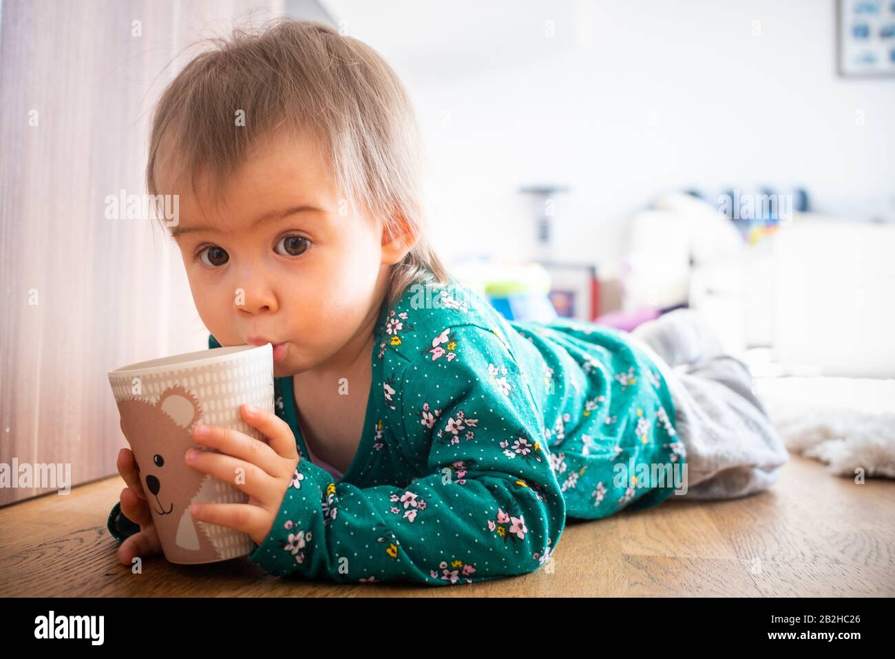 12-15th month old adorable caucasian girl lie on wooden floor with cup in hands. Stock Photo