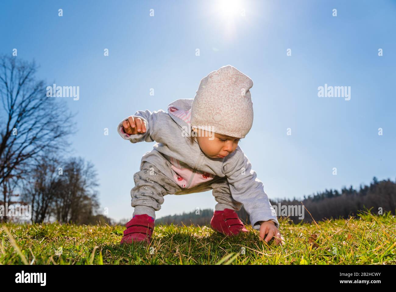 Baby wearing warm beanie hat, sweatshirt and red boots outdoors in rural area discovering nature in spring, sunny day. Stock Photo