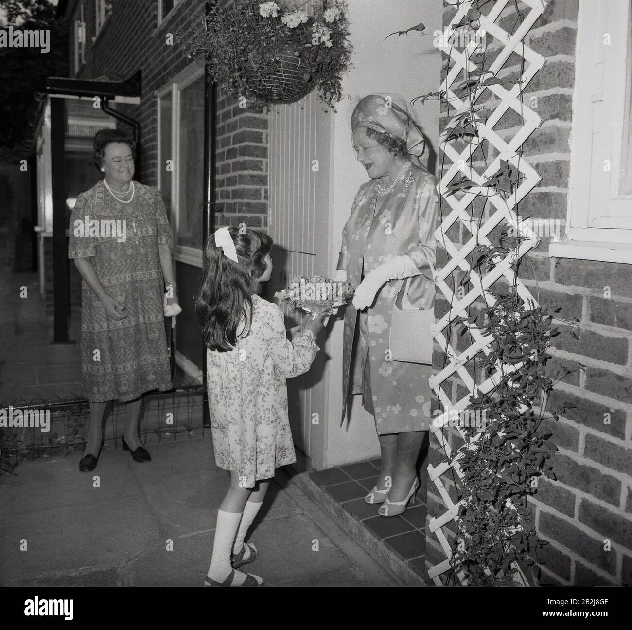 1970s, historical, a young girl presenting a flower bouquet to the the Queen mother standing in a doorway of a house as she visits a street in South London, England, UK. Stock Photo
