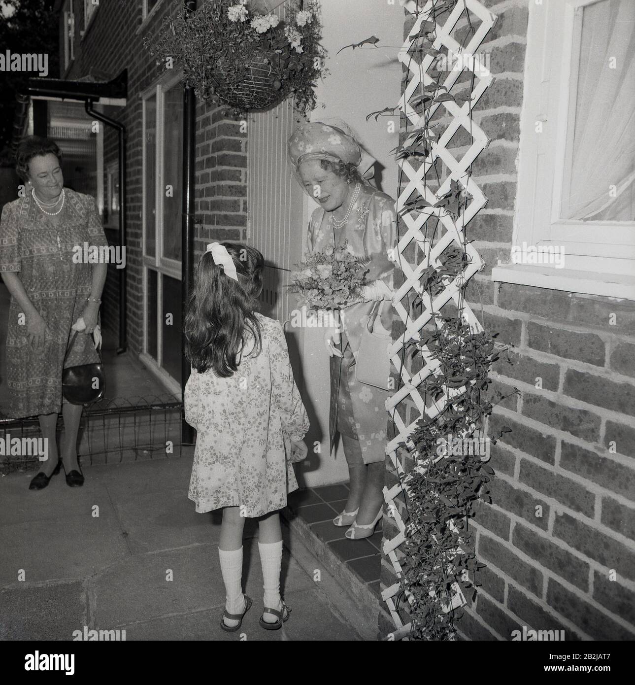 1970s, historical, a young girl presenting a flower bouquet to the the Queen mother standing in a doorway of a house as she visits a suburb in South London, England, UK. Stock Photo