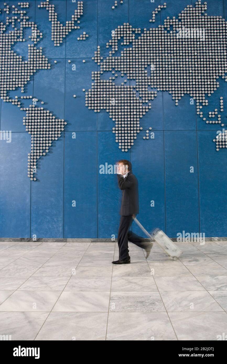 Business man with suitcase using mobile phone while walking past world map on wall Stock Photo