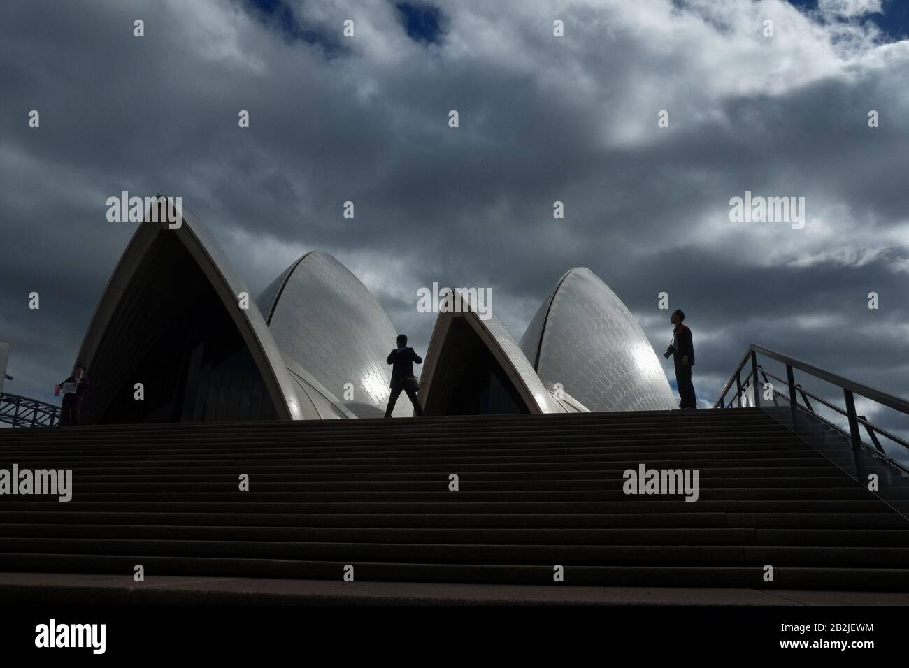 Silhouette of two men against the white sails and dramatic storm cloud sky at the top of the monumental steps, upper podium of the Sydney Opera House Stock Photo