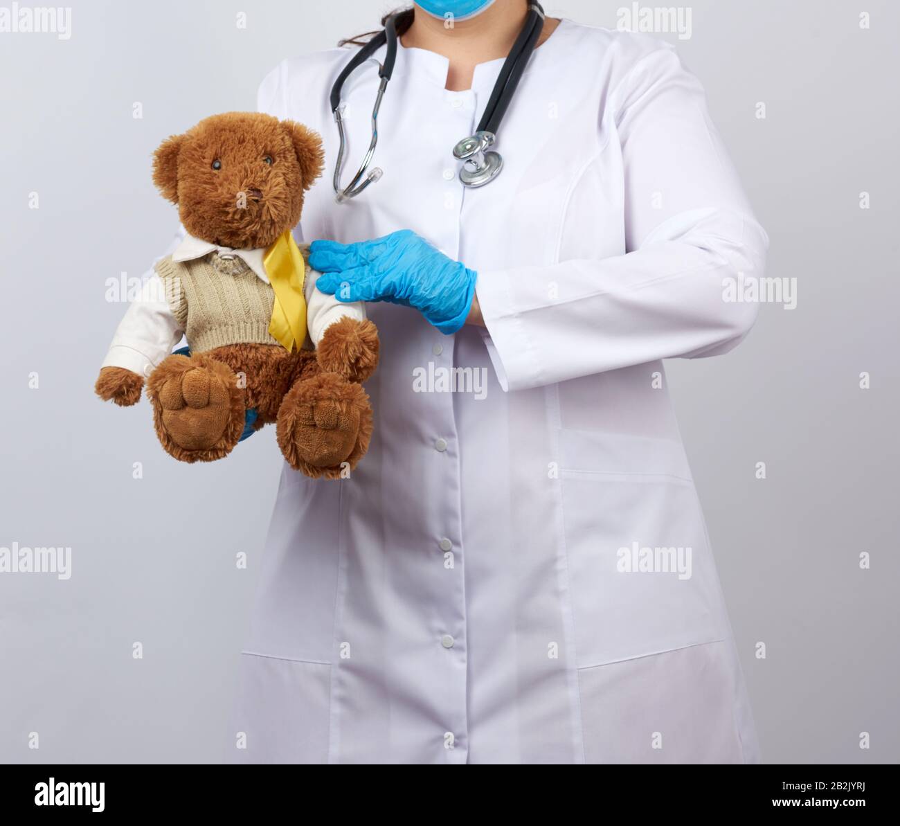 pediatrician in white coat, blue latex gloves holds a brown teddy bear with a yellow ribbon on a sweater, concept of the fight against childhood cance Stock Photo