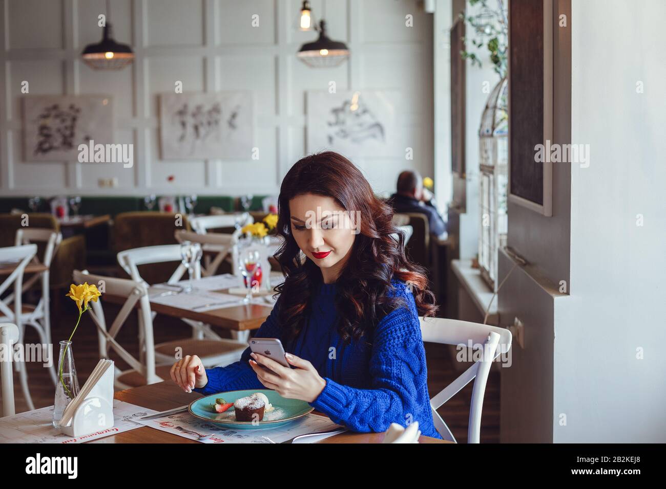 the woman in the cafe sits at a table, drinks coffee and looks at window. A woman with red lipstick and in a blue sweater is waiting for a meeting, ta Stock Photo