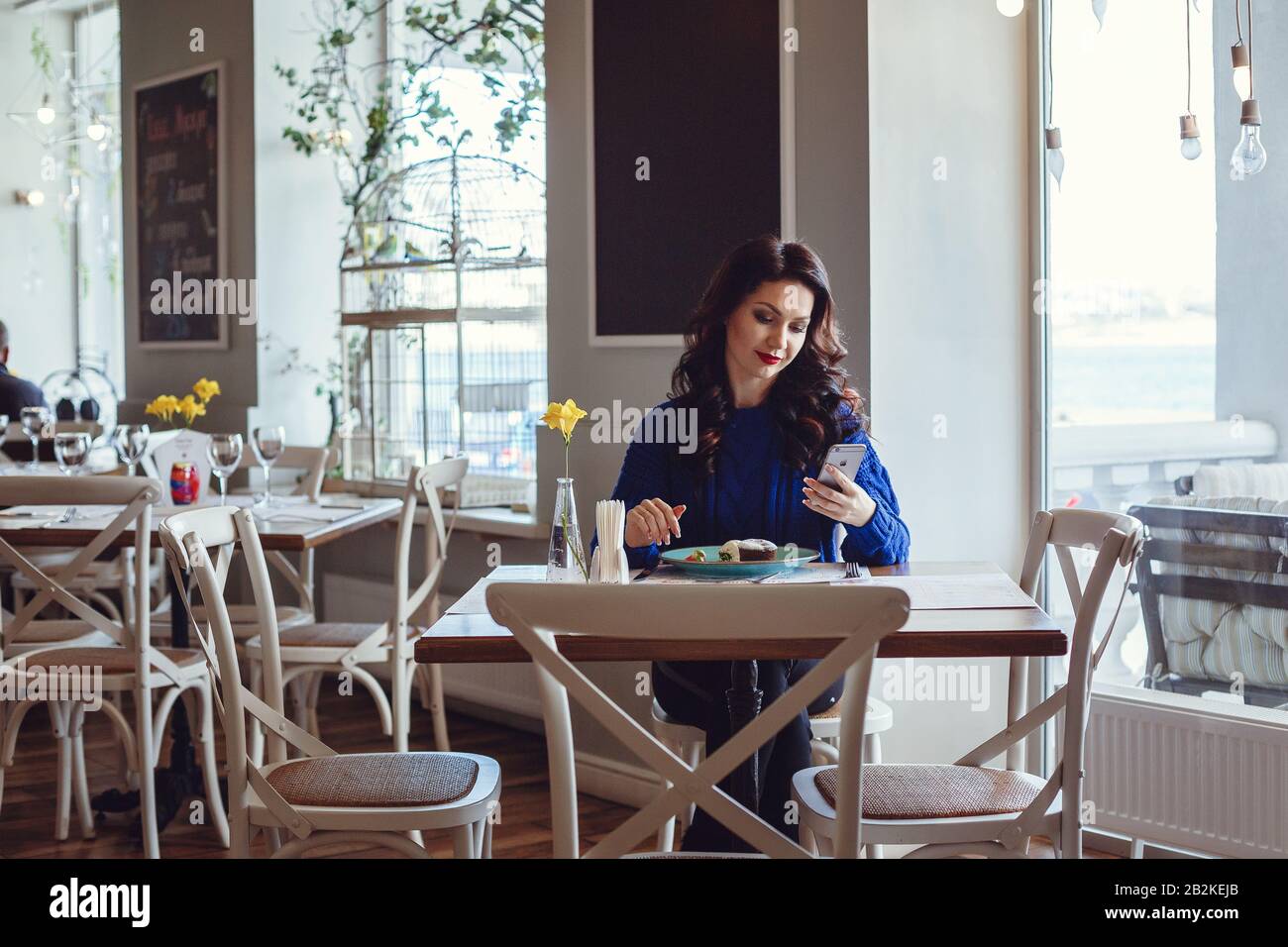 the woman in the cafe sits at a table, drinks coffee and looks at window. A woman with red lipstick and in a blue sweater is waiting for a meeting, ta Stock Photo