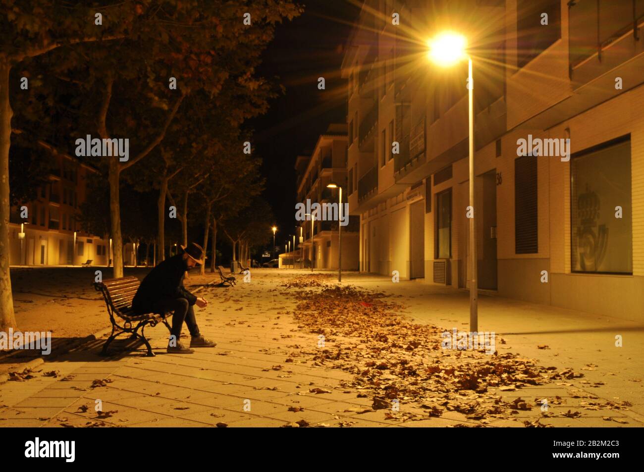 Lonely man sitting on a urban bench under the light of a streetlight and dry leaves all over the ground/ Book cover variety Stock Photo