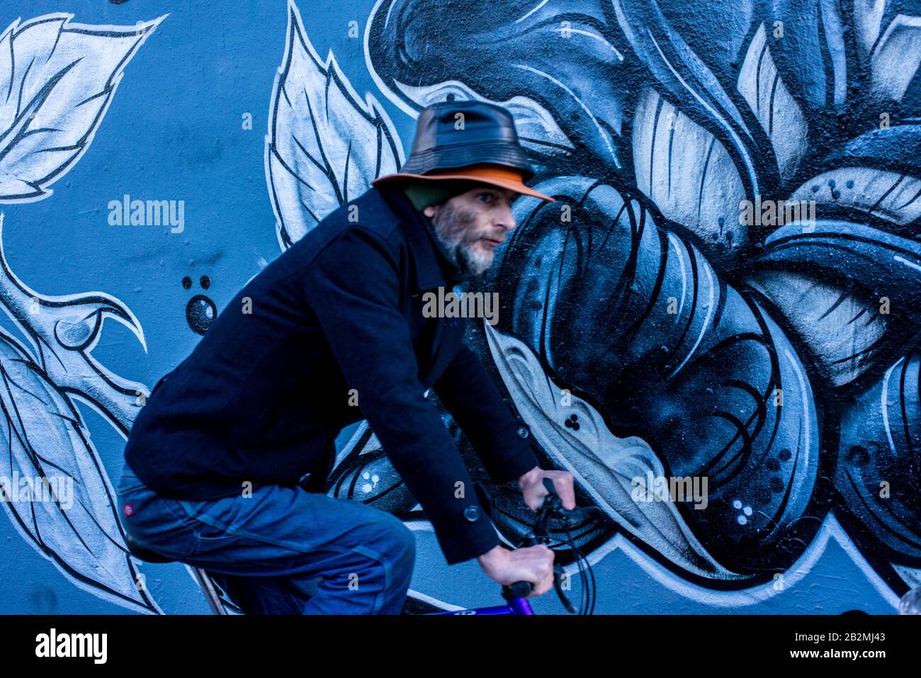 A man dressed in blue cycles past a piece of blue flower-patterned piece of graffiti art while wearing two hats on top of each other. Stock Photo