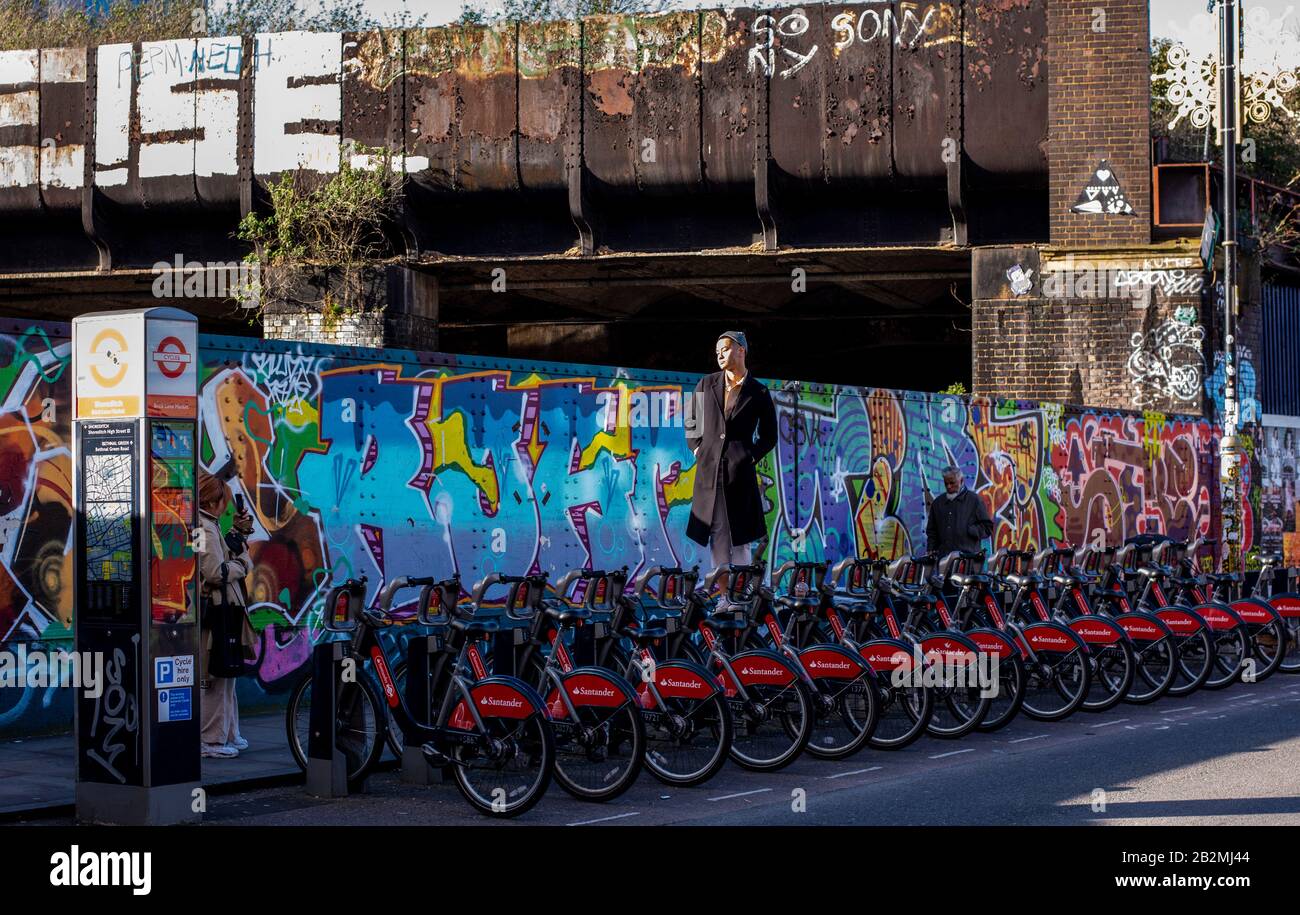 A young Asian man stands on a bicycle in the middle of a row of parked bikes for hire, to have his photo taken in Brick Lane, London. Stock Photo