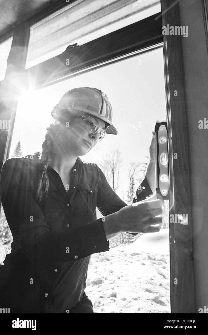 close up on female construction inspector uses spirit level on vertical surface next to a window with sun in the sky view. vertical, black and white. Stock Photo