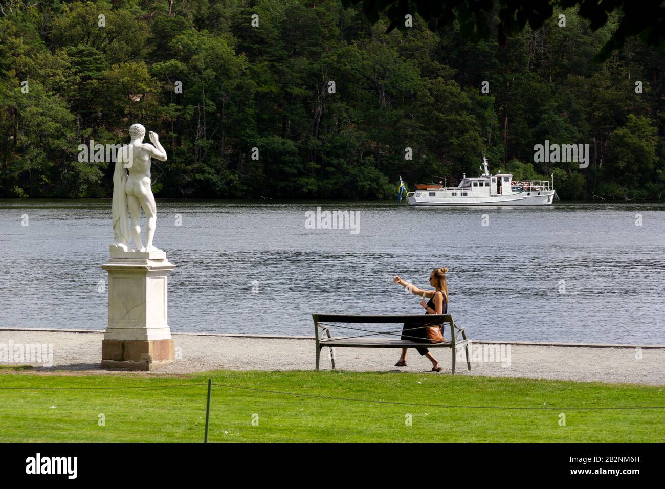 Stockholm, Sweden - July 13, 2019: A woman sitting on a bench next to a statue and water taking a selfie in the wonderful Drottningholm. Stock Photo
