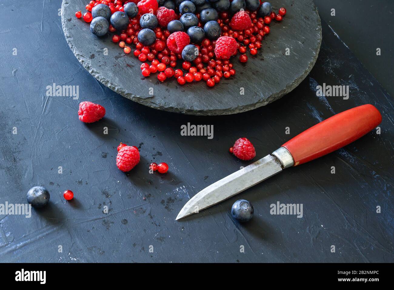 Fresh blueberries, raspberries and redcurrants, kitchen knife, black dish, dark backdrop Stock Photo