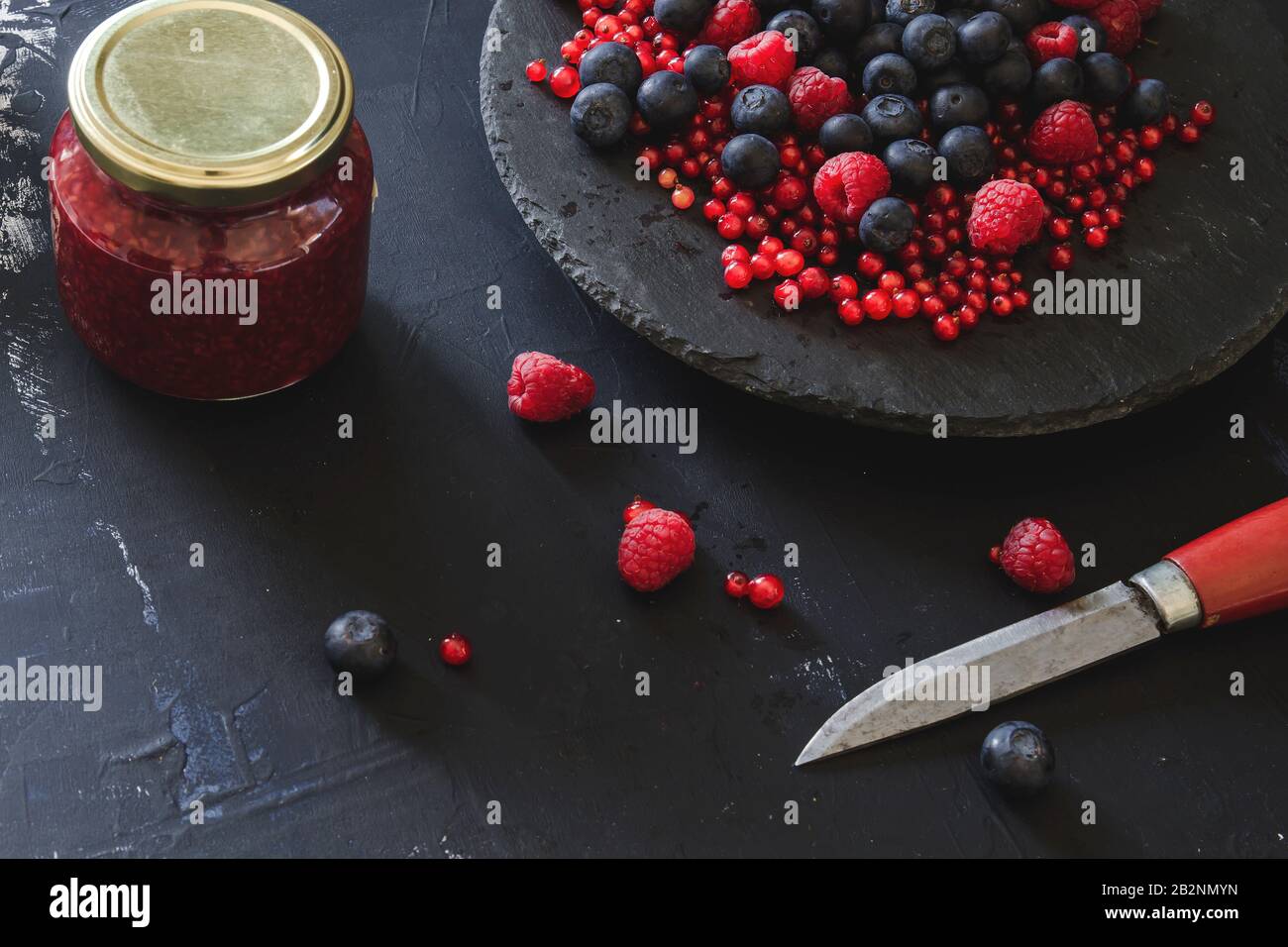 Homemade jam with fresh blueberries, raspberries and redcurrants, kitchen knife, black dish, dark backdrop Stock Photo