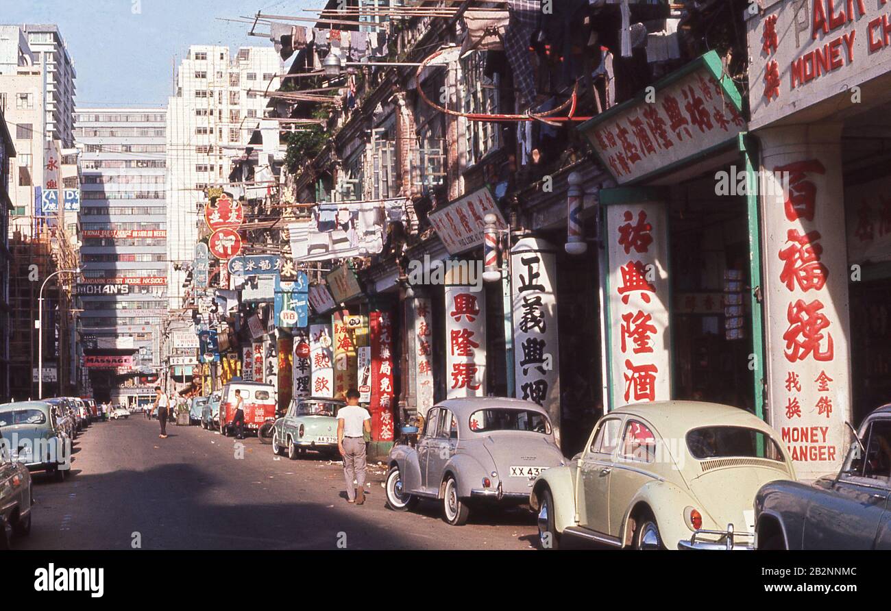 1960s, historical, daytime and a street in the old town, Hong Kong, showing retail outlets, chinese lettering on the support pillars of the pavement walkway and motorcars of the era parked up, including a British made Morris Minor and a Volkswagen Beetle. Stock Photo