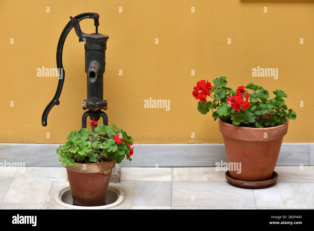 Old water pump with two pots of geraniums in a patio with marble floor and yellow wall Stock Photo