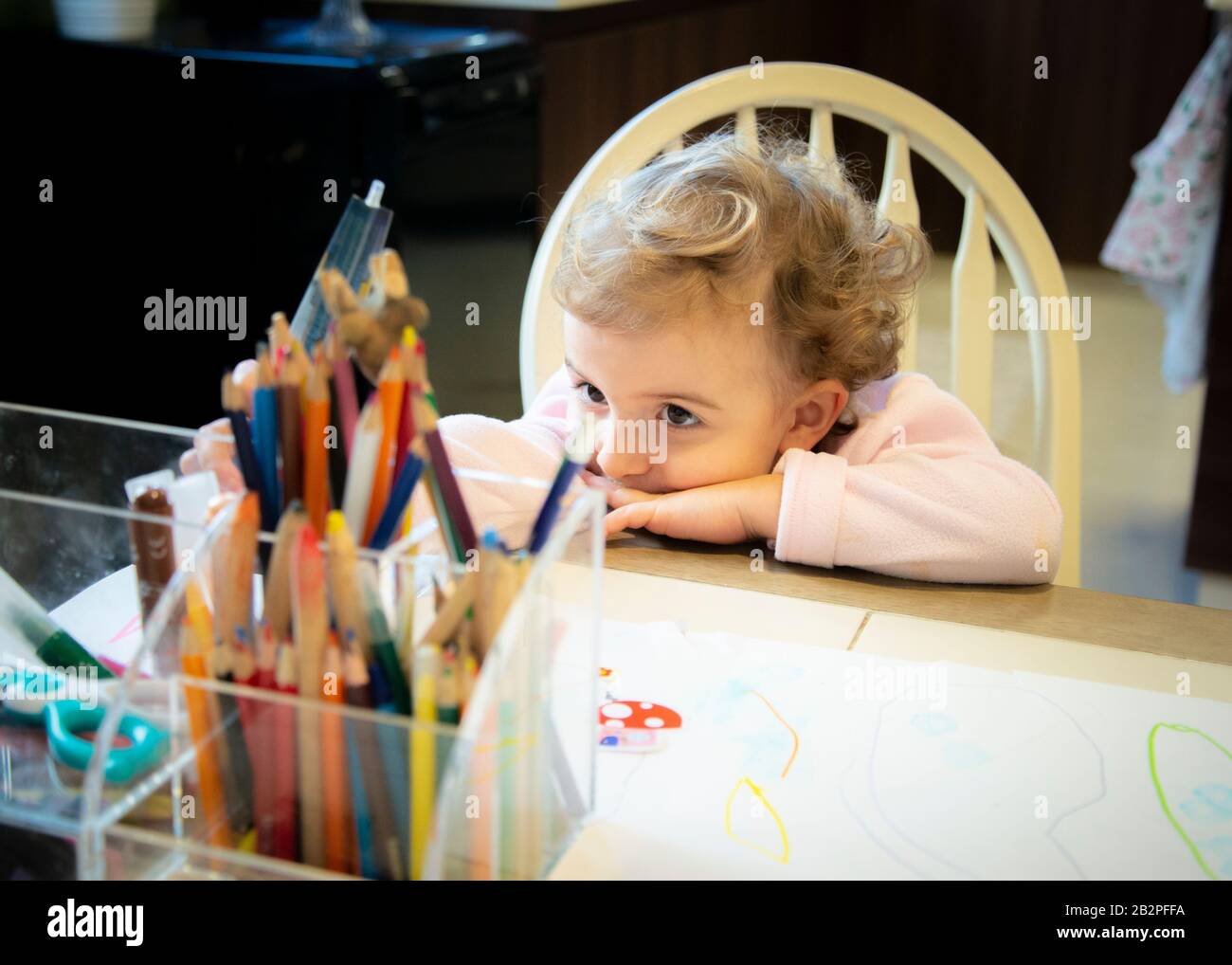 child leaning on table looking thoughtful,staring at holder containing colored,pencils and crayons. Stock Photo