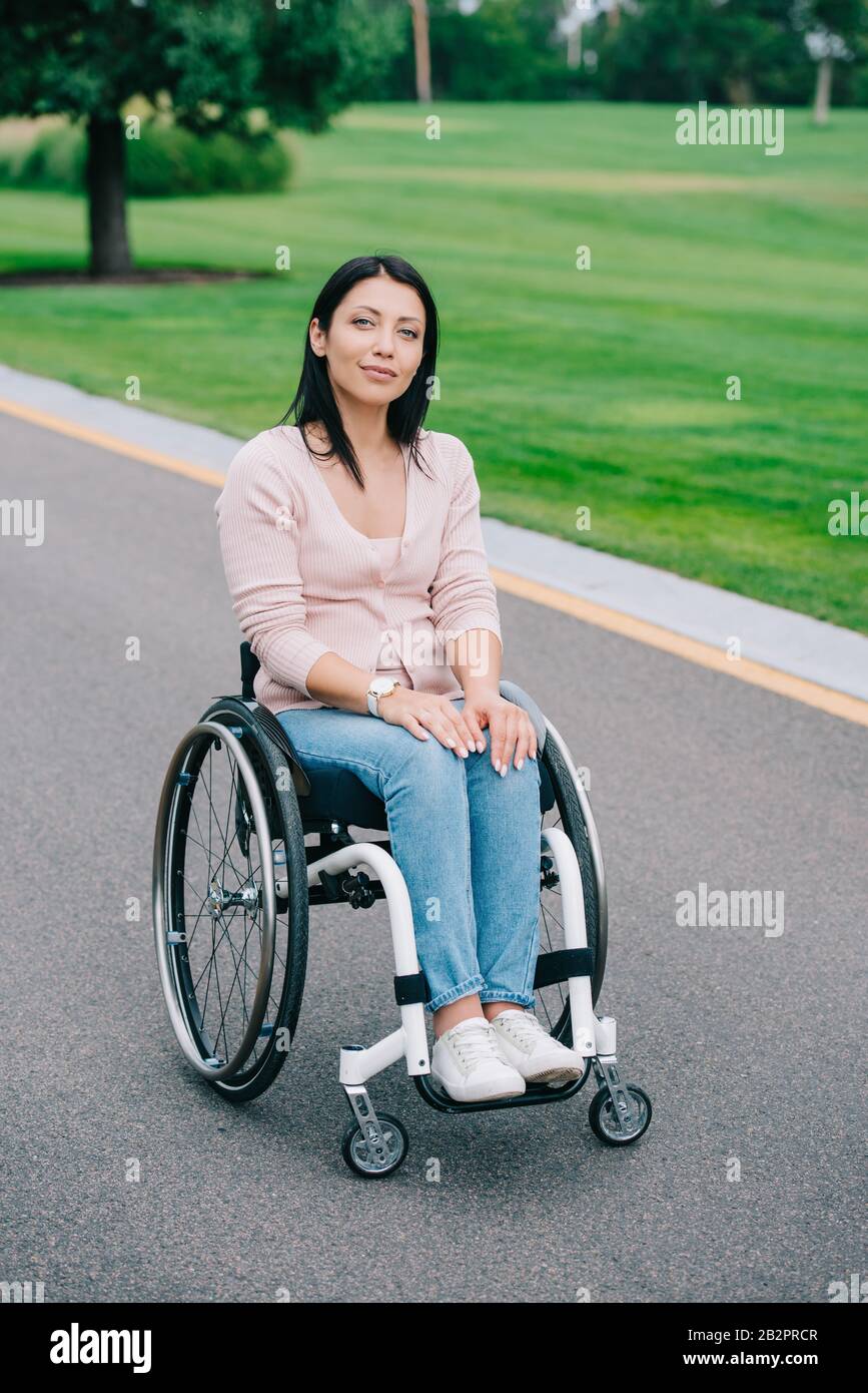 young disabled woman in wheelchair smiling at camera while walking in ...