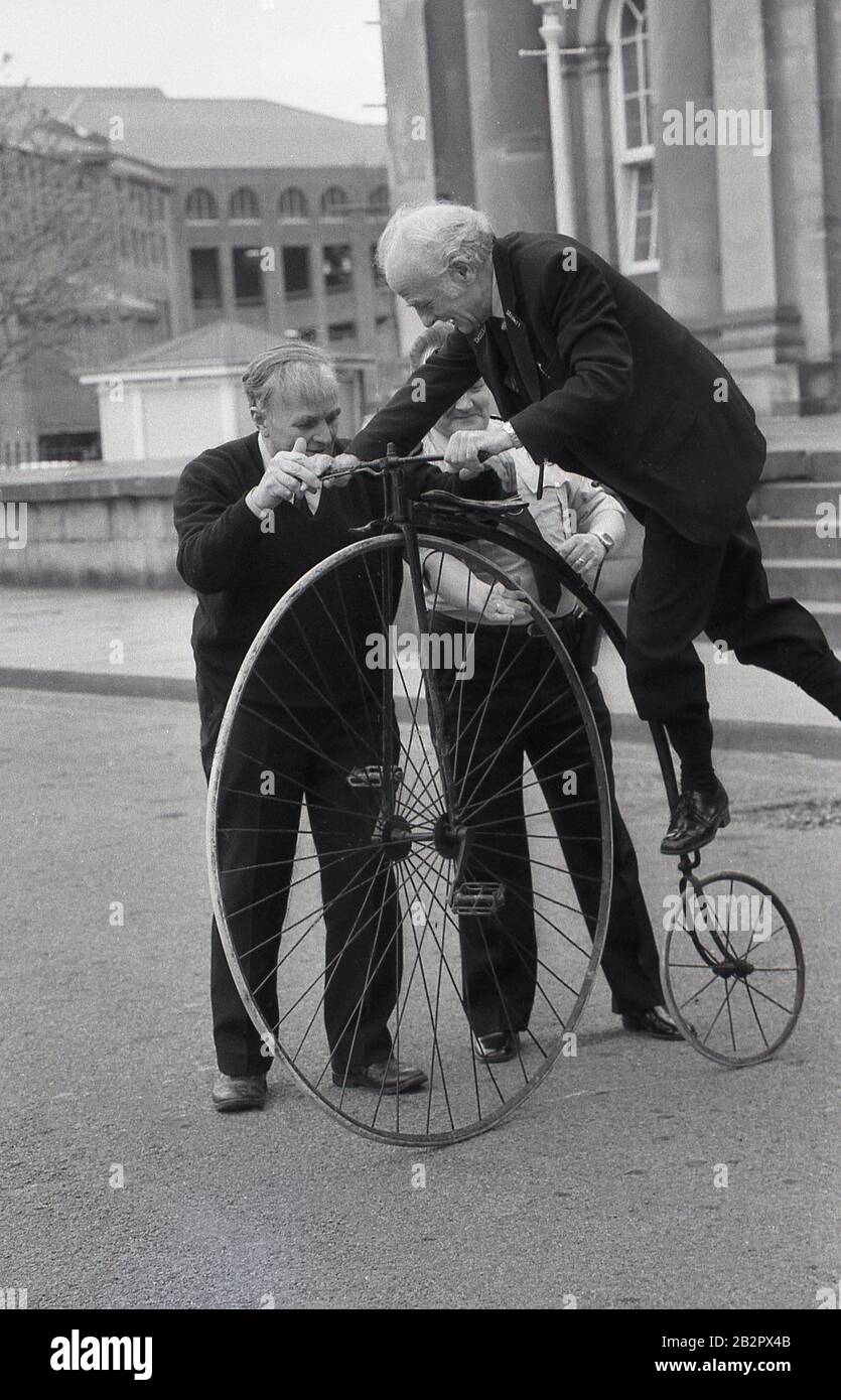 1980s, historical, an elderly man attempting to get on or mount a penny-farthing bicycle, also known as a high wheeler or an 'ordinary', York, England. The machine was the first to be known as a 'bicycle' and with its large front wheel was capable of high speeds. However it was difficult to mount, ride and there was a danger of falling of and it was superceded by the modern 'safety' bicycle. Stock Photo