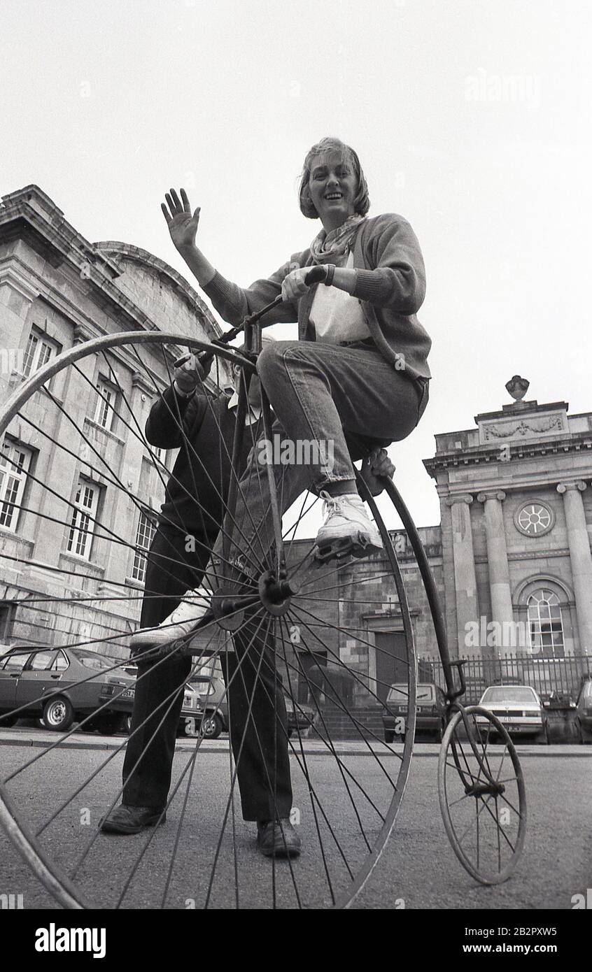 1980s, historical, a woman sitting on a penny-farthing bicycle, also known as a high wheeler or an ordinary. Popular in the 1870s and 80s, with its large front wheel and small back wheel, it was the first machine to be known as a 'bicycle'. Difficult to mount and to ride and with a danger of falling off, given the height of the front wheel, it was superceded by the modern 'safety bicycle'. Stock Photo