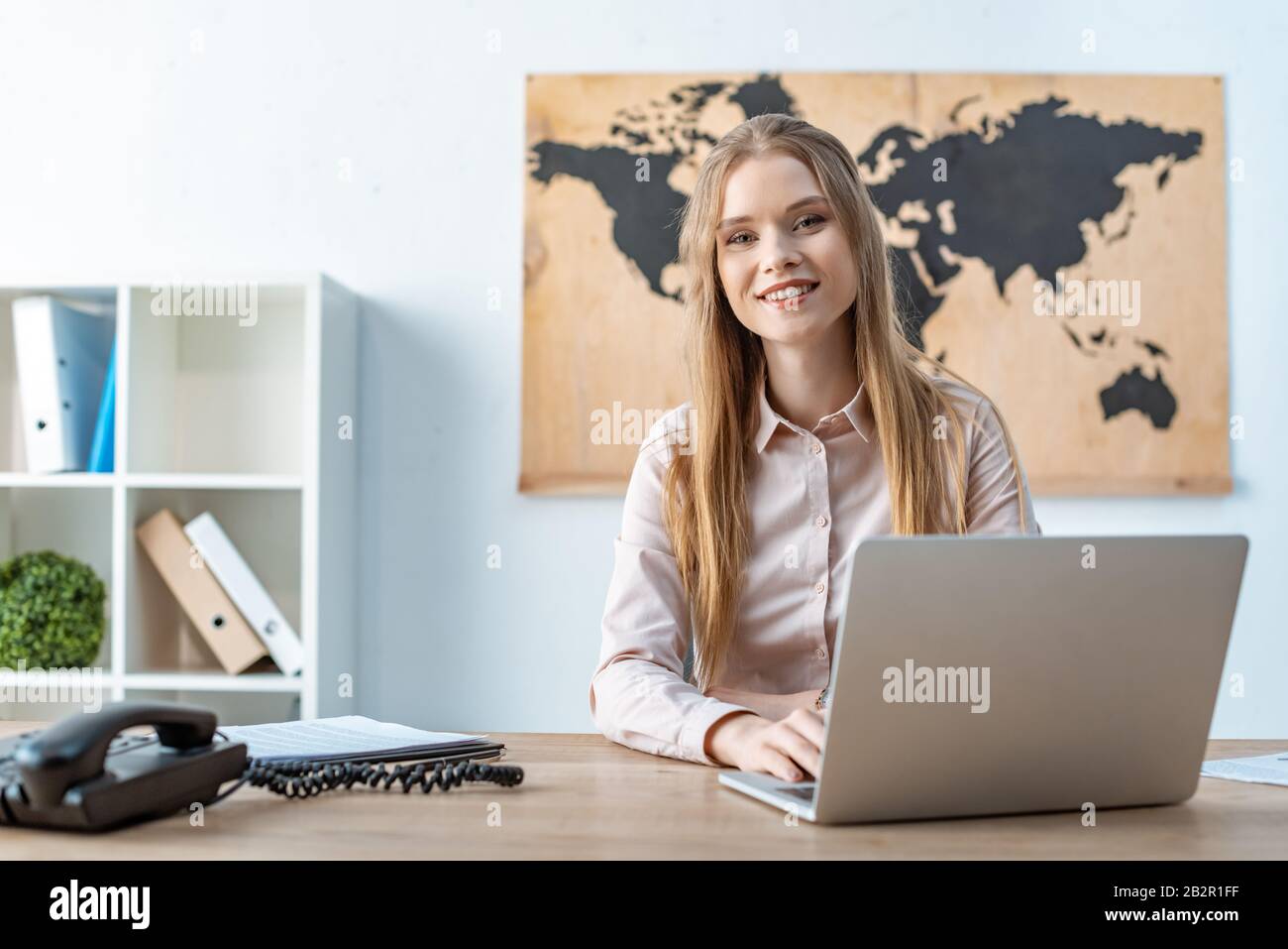 attractive travel agent smiling at camera while using laptop Stock Photo