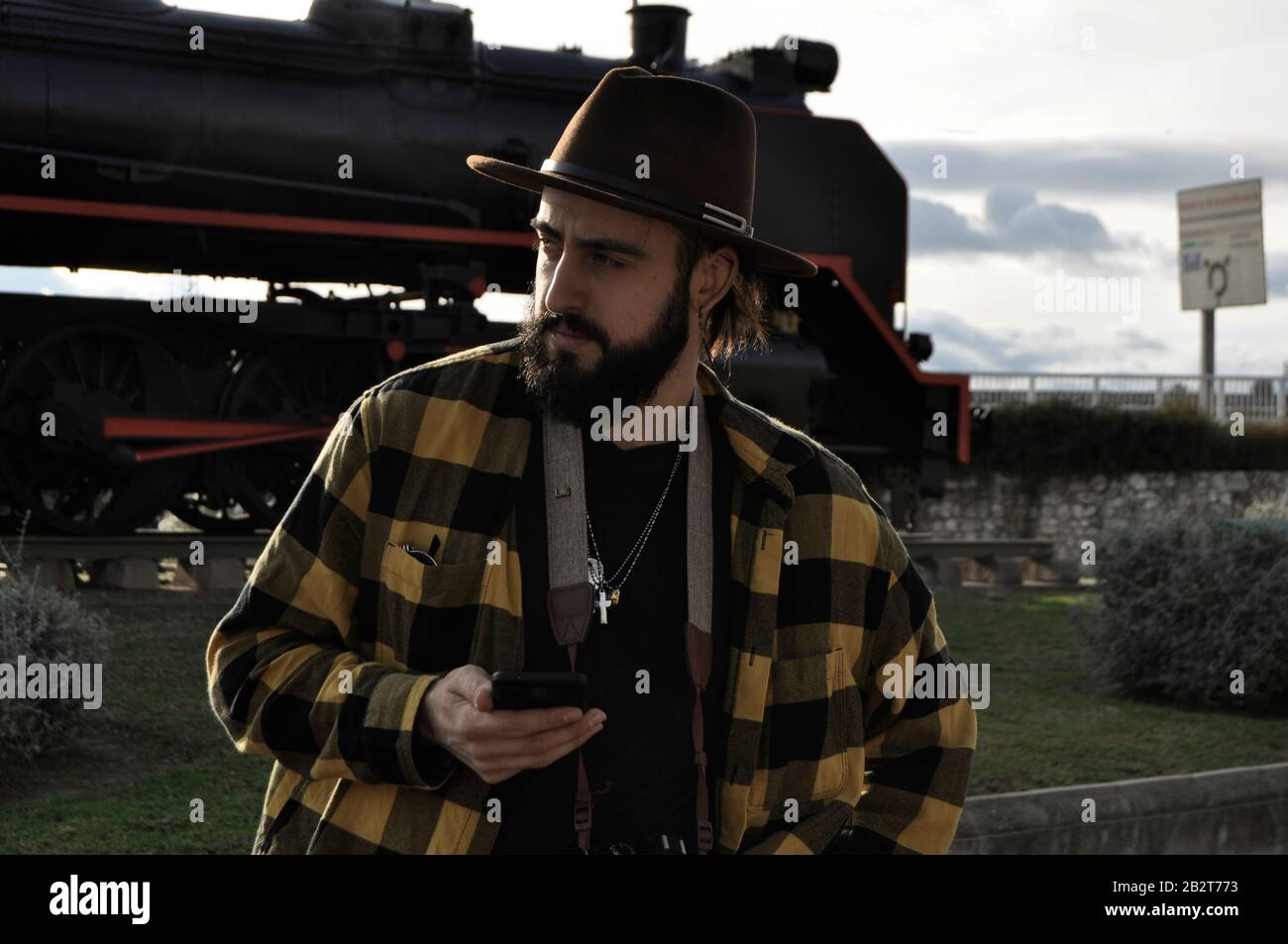 Bearded man wearing a cowboy hat, next to a train station using a smartphone/ Young man wearing a 1950 fedora hat Stock Photo