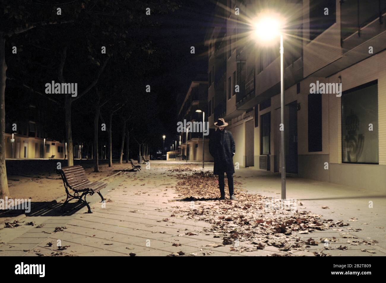 man wearing suit and hat on a Spanish street with his head down under the light of a lantern and dry leaves all over the ground/ Abstract imagery Stock Photo