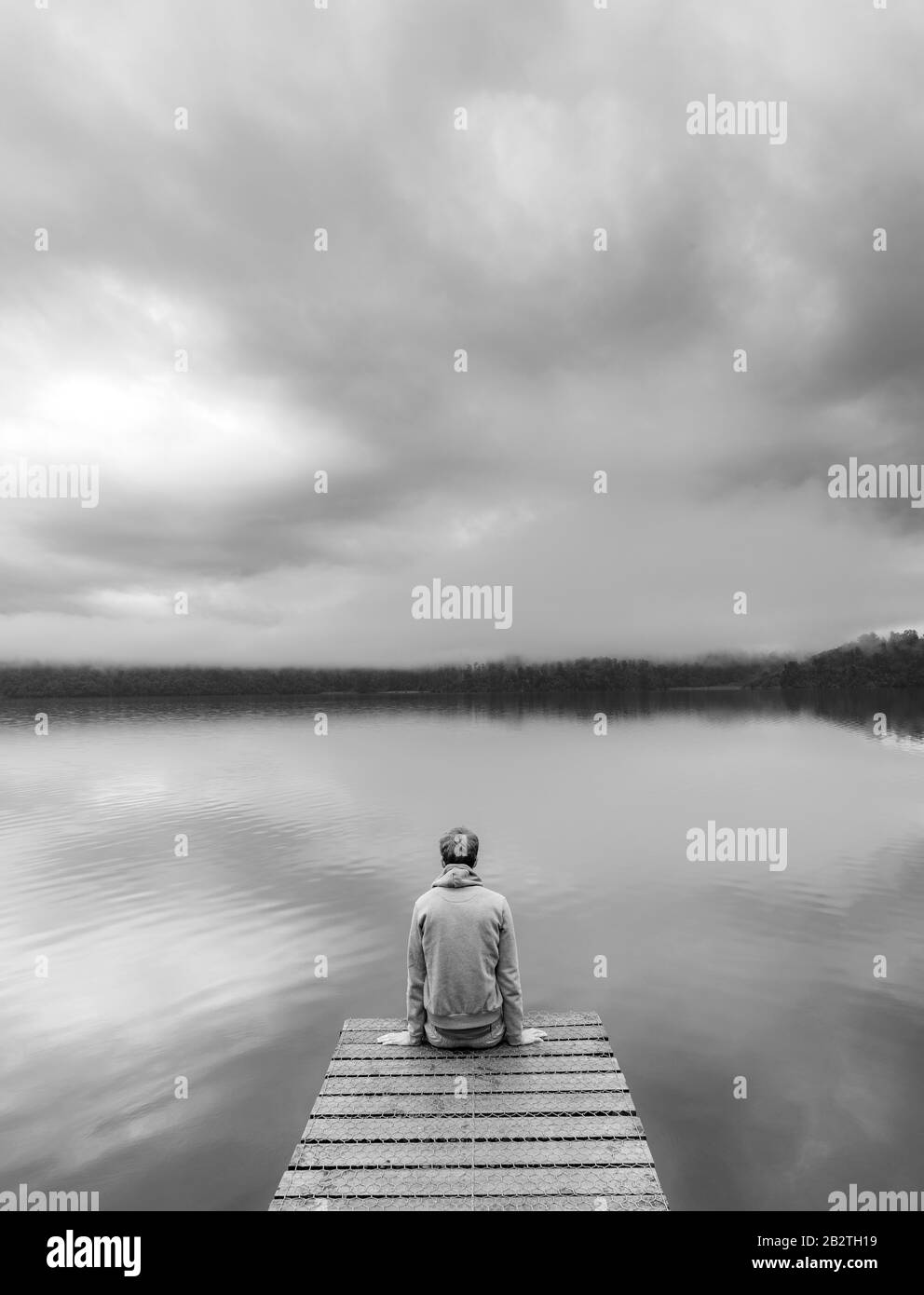 Young man sitting on a jetty looking over a lake, foggy atmosphere, Lake Mapourika, West Coast, South Island, New Zealand Stock Photo