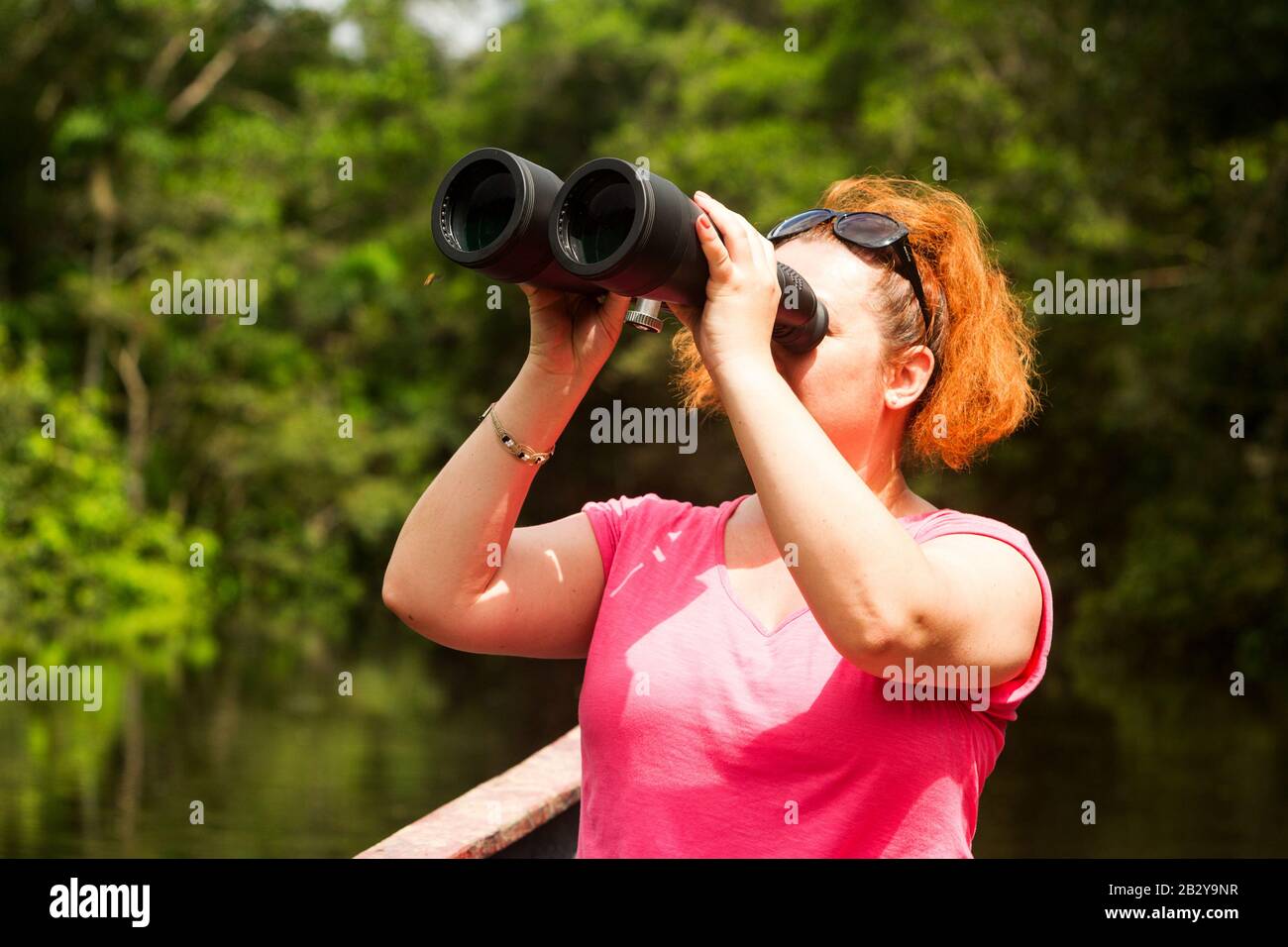 Tourist Woman With High Power Binoculars In Amazonian Jungle Against Dense Vegetation Stock Photo