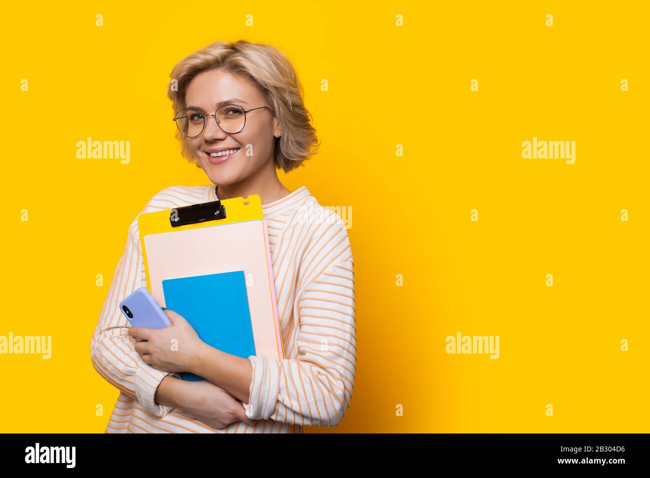 Stunning blonde teacher looking through eyeglasses while holding some books and posing on a yellow wall with freespace Stock Photo