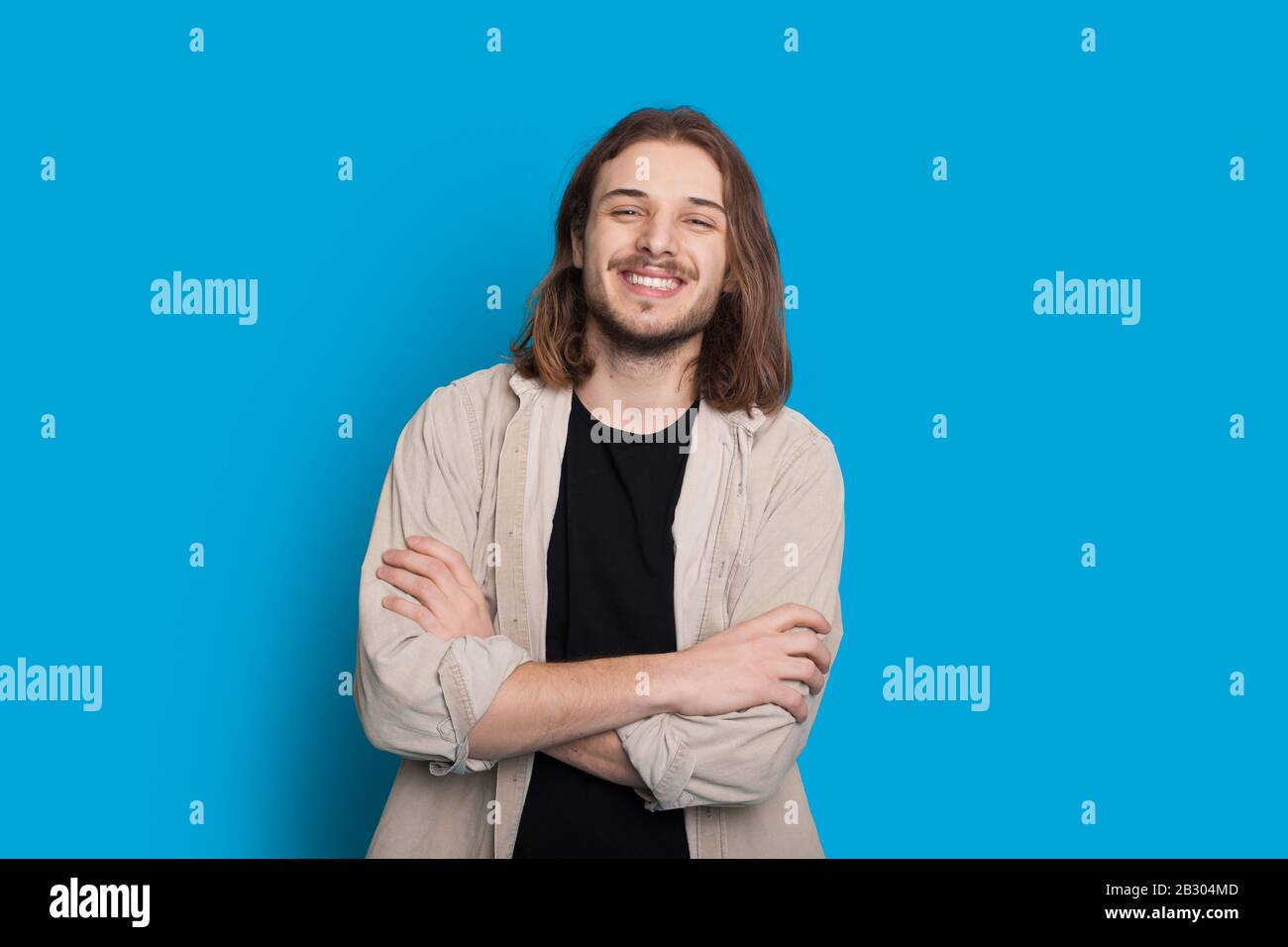 Confident caucasian man with long brown hair and beard is posing with crossed hands on a blue wall Stock Photo