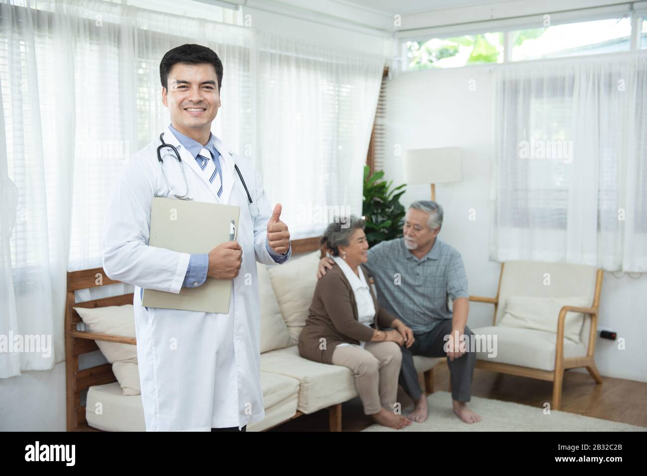 Young handsome caucasian male doctor standing hand holding document file look at camera with smile and two elderly old senior asian couple sit on sofa Stock Photo