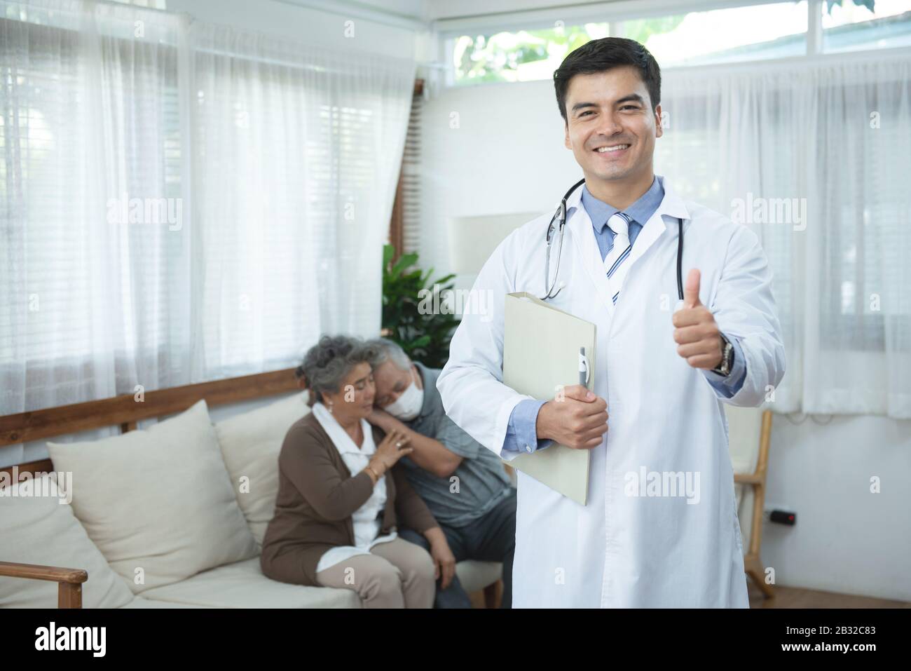 Young handsome caucasian male doctor standing hand holding document file look at camera with smile and two elderly old senior asian couple sit on sofa Stock Photo