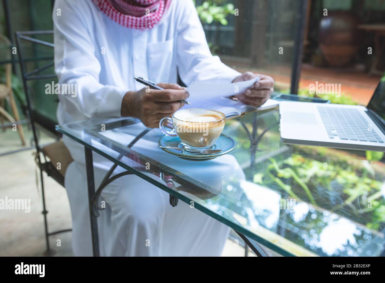 Young handsome businessman arab muslims in coffee shop wearing hijab dressed  write on book file with laptop on table.focus on glass of hot coffee. Stock Photo