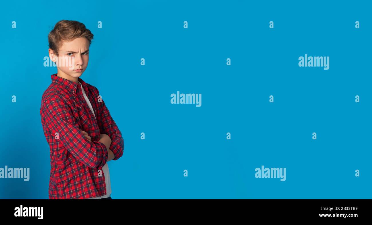 Frowning Grumpy Teen Boy Standing With Folded Arms Over Blue Background Stock Photo
