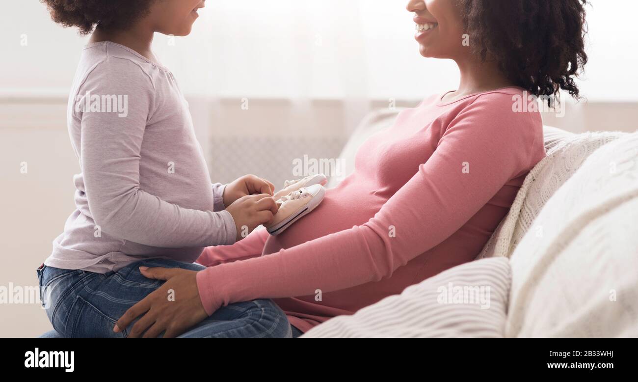 Little Daughter Putting Small Baby Shoes At Pregnant Afro Mother's Belly Stock Photo