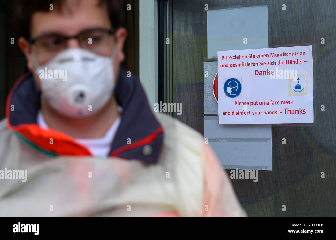 04 March 2020, Baden-Wuerttemberg, Stuttgart: A volunteer employee of the German Red Cross (DRK) is standing in front of a sign in front of the corona outpatient clinic of the Klinikum Stuttgart in the Katharinenhospital saying 'Please put on a mouthguard and disinfect your hands'. Patients can have themselves tested in the Corona Outpatient Clinic. Photo: Sebastian Gollnow/dpa Stock Photo
