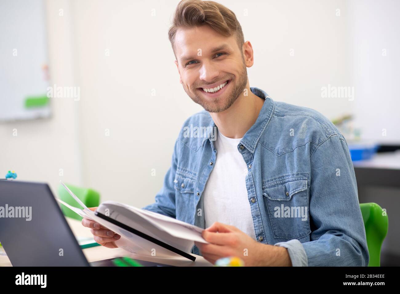 Male teacher sitting at desk, holding papers, smiling Stock Photo - Alamy