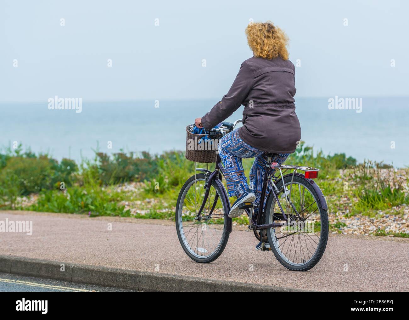 Woman riding a bicycle along a seafront pavement by the sea. Female cyclist riding a bike in the UK. Stock Photo